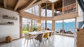 Une salle à manger spacieuse, dotée d’une grande table et de chaises, de poutres en bois et de grandes fenêtres offrant une vue sur les montagnes.