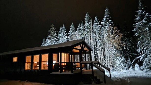 Eine Hütte mit Veranda und großen Fenstern, umgeben von schneebedeckten Bäumen.