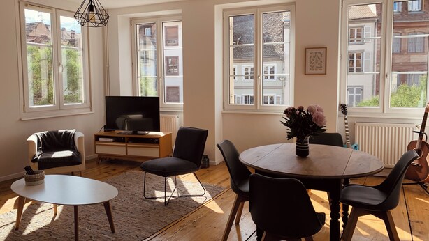 Een woonkamer met een televisie, een eettafel met stoelen, een bank en een raam met uitzicht op gebouwen.