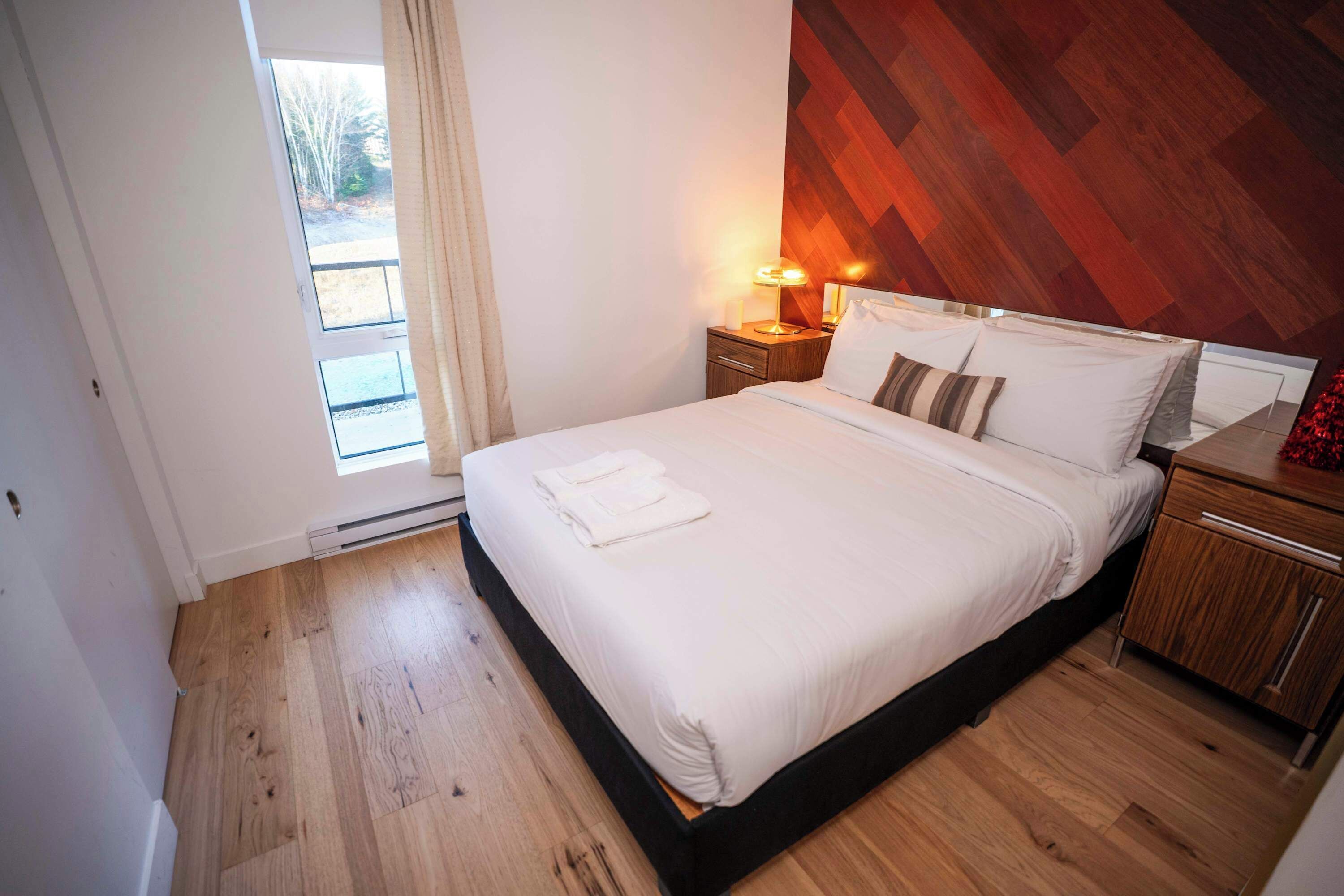 Una cama bien hecha con sábanas blancas, un cabecero de madera, una mesita de noche con una lámpara y vistas al exterior a través de una puerta.