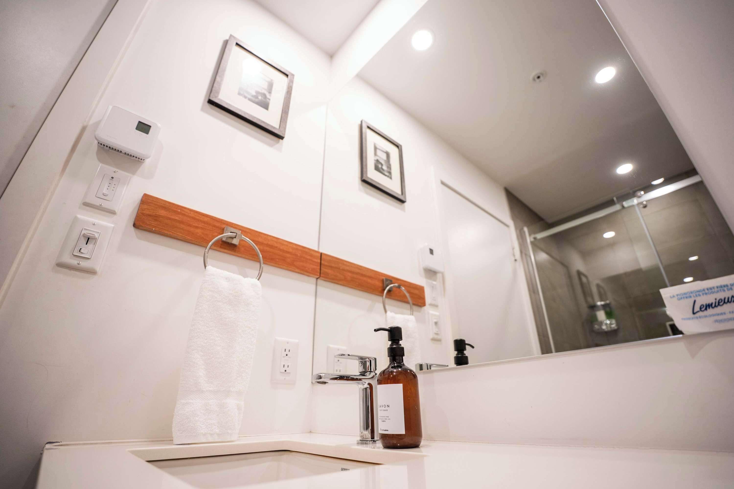 A modern bathroom with a white sink, a mirror, and a wooden panel above the mirror. There are two framed pictures on the wall, a towel hanging on the towel rack, and a bottle of soap on the sink.