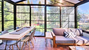 Ein Wintergarten mit Holztisch, Stühlen und einer Couch, mit Blick auf eine bewaldete Landschaft.