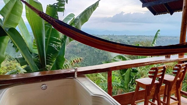 Una terraza de madera con un jacuzzi, sillas de madera y una hamaca con vistas a un paisaje exuberante.