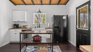 A kitchen with white cabinets, a black refrigerator, a wooden table with a red pot, and a window with a view of trees.