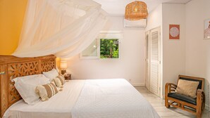 A bedroom with a canopy bed, a wooden headboard, a bedside table with a lamp, a chair, and a window with a view of greenery. - Alizéa (Le Diamant)