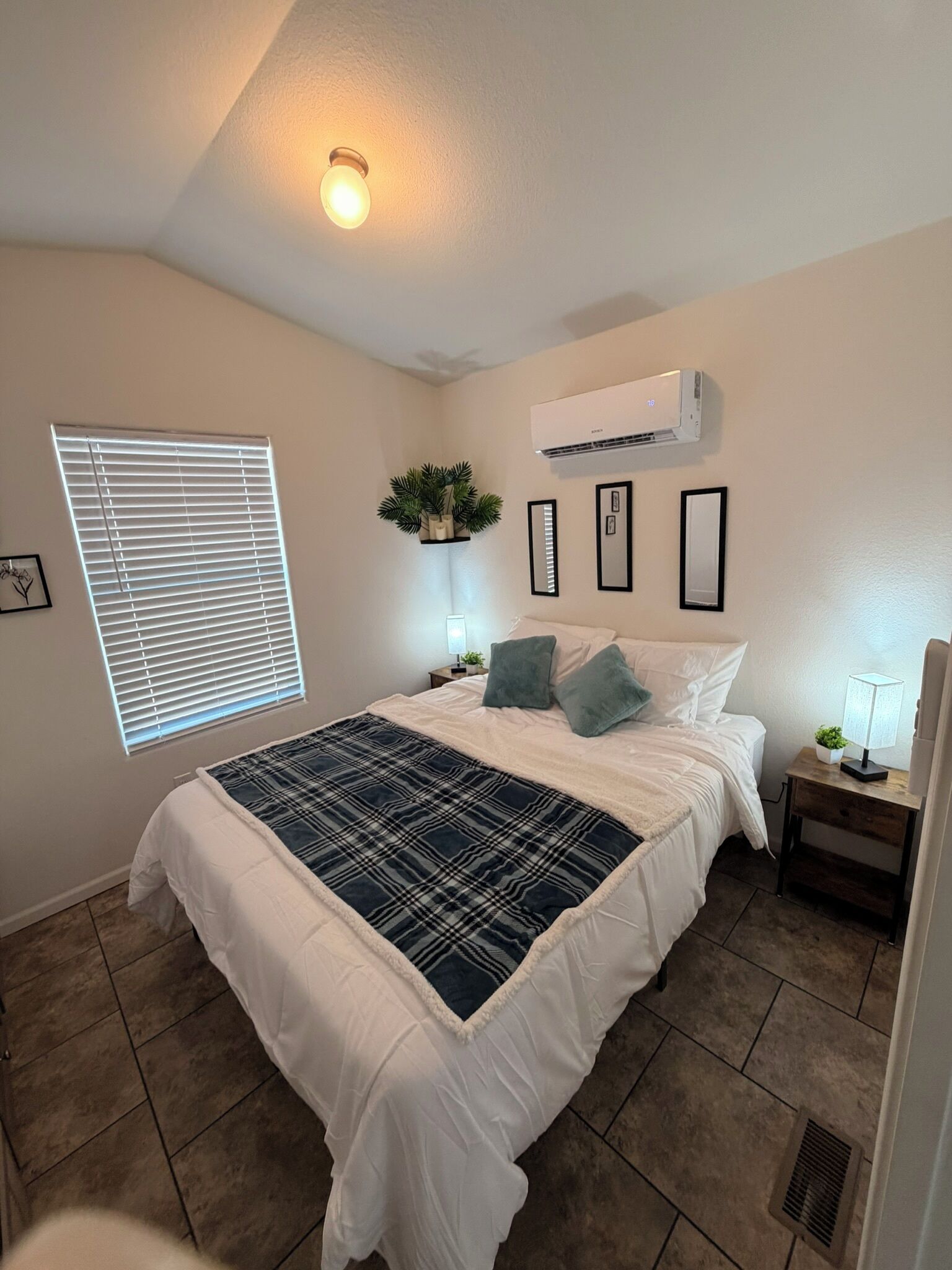 Un dormitorio con cama, ventana con persianas, aire acondicionado, mesita de noche con lámpara y una planta pequeña en la pared.