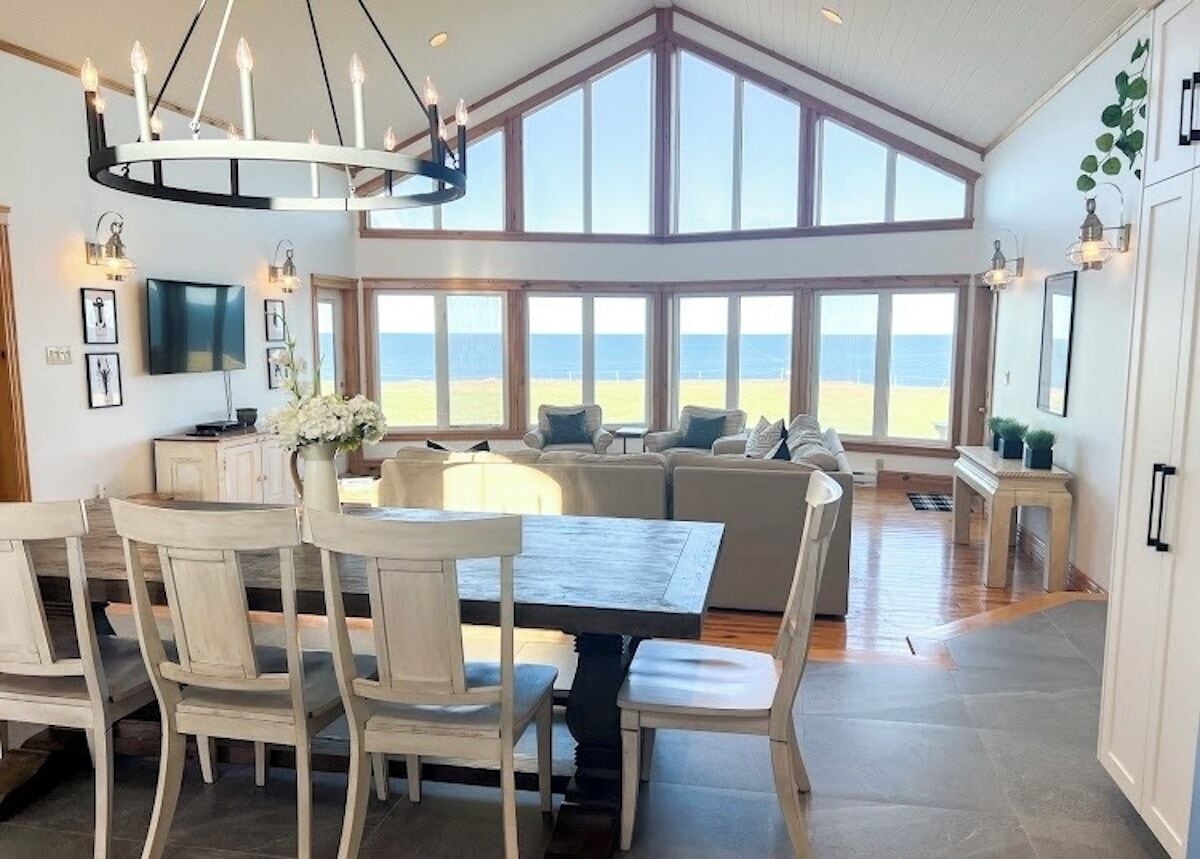 A spacious living area with a large window overlooking the beach, a dining table with chairs, and a modern chandelier.