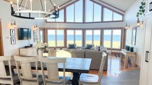 A spacious living area with a large window overlooking the beach, a dining table with chairs, and a modern chandelier. - Ocean Front Cottage With Panoramic Water Views- Seconds To Cousin's Shore Beach (Kensington)