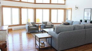 Un vaste espace de vie comprenant un grand canapé d’angle gris, deux fauteuils, une table basse et une petite table d’appoint. La pièce est dotée de grandes fenêtres et d’un plafond haut.