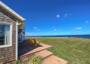 A house with a deck and chairs, a grassy lawn, and a view of the ocean. - Ocean Front Cottage With Panoramic Water Views- Seconds To Cousin's Shore Beach (Kensington)