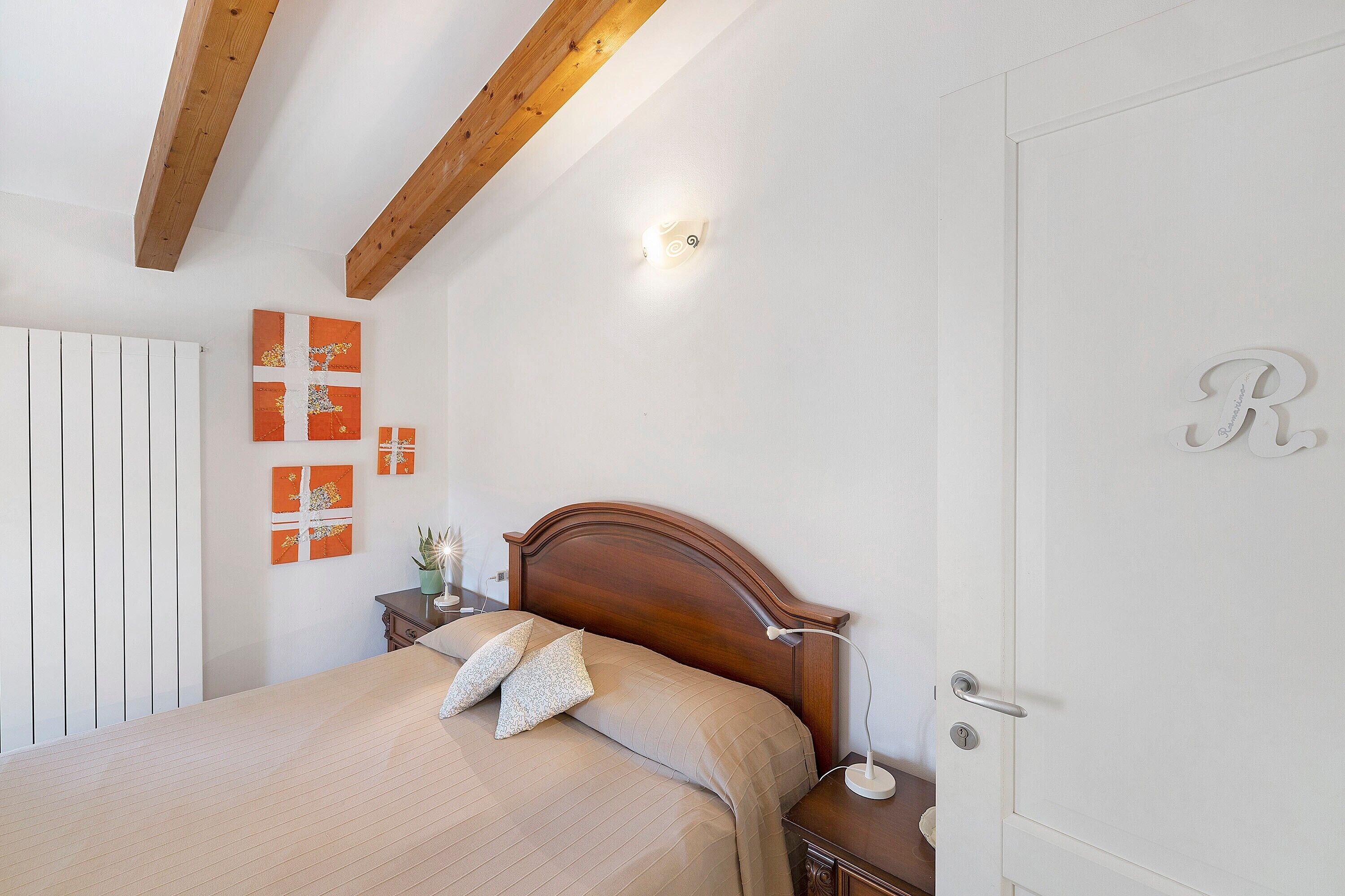 Un dormitorio con cabecera de madera, cama con ropa de cama color beige, mesita de noche con lámpara y una puerta con la letra 'R'.