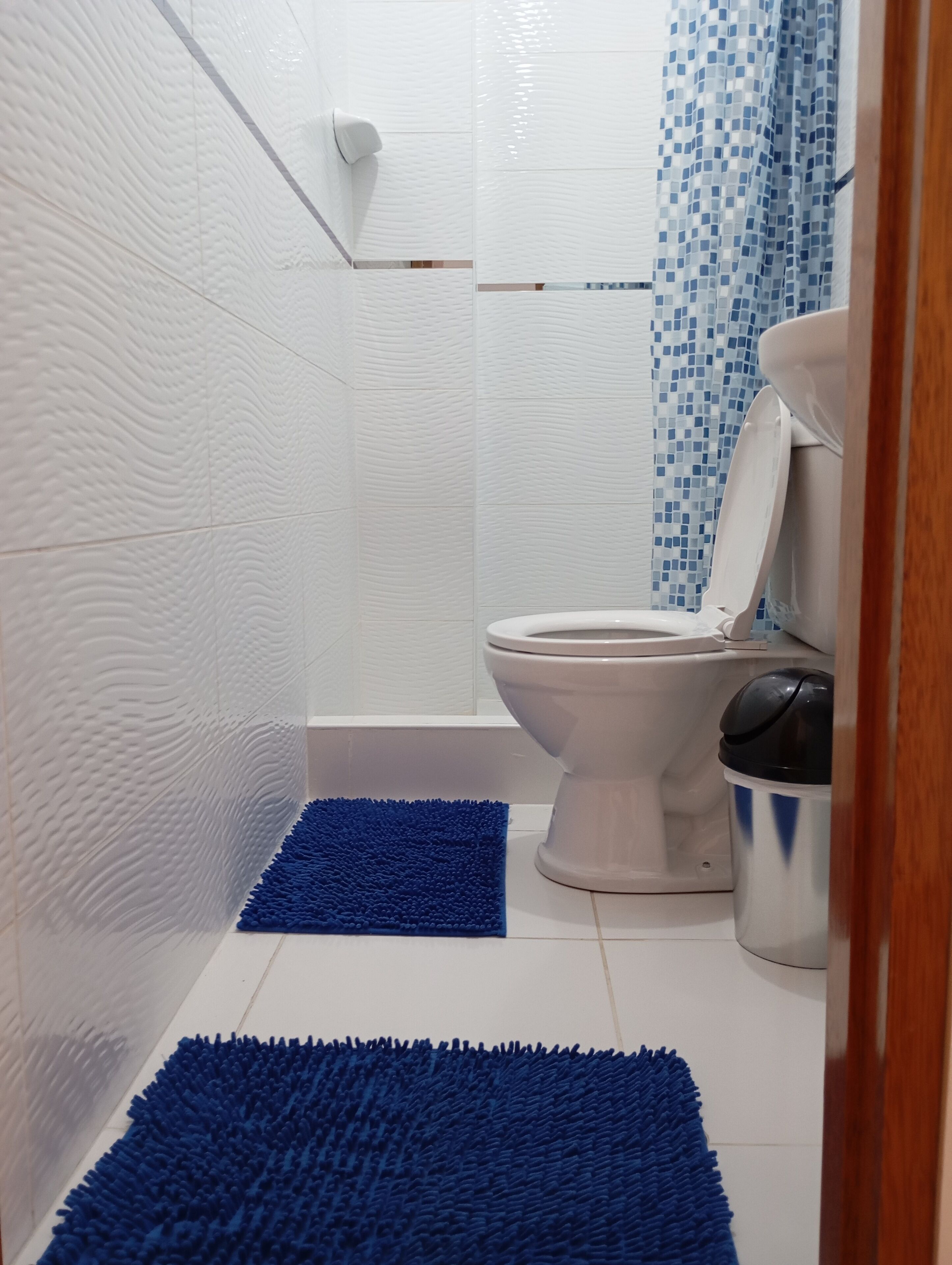 Un baño con paredes blancas con textura, un inodoro blanco, una cortina de ducha azul y un tapete de baño azul.