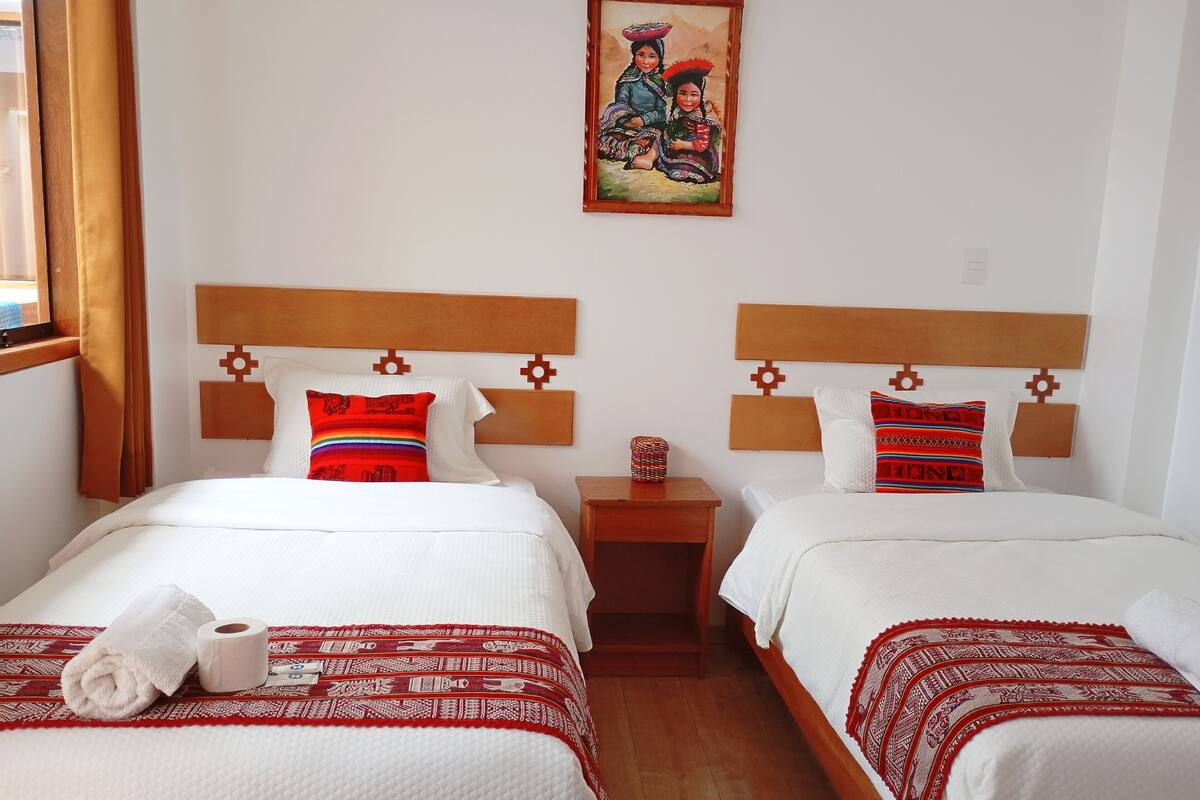 Habitación de hotel con dos camas, cada una con cabecero de madera, ropa de cama blanca y mantas rojas estampadas. Un cuadro cuelga de la pared sobre las camas.