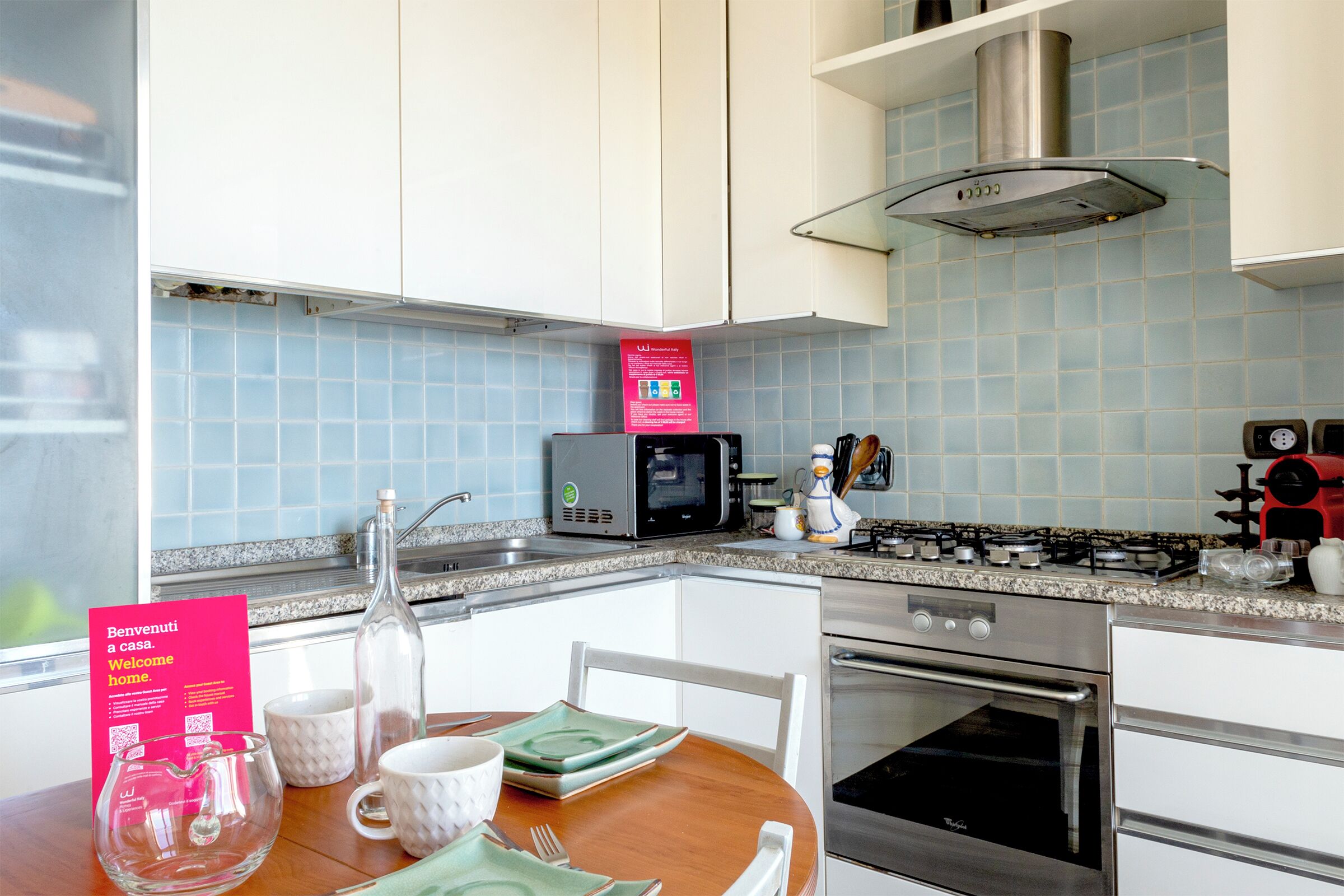A modern kitchen with white cabinets, a microwave, a gas stove, and a stainless steel oven. There is a dining table with plates, cups, and a bottle.