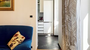 Ein Wohnzimmer mit einem blauen Sofa und einem gemusterten Kissen, das in eine Küche mit weißen Schränken und einem Fliesenboden übergeht.