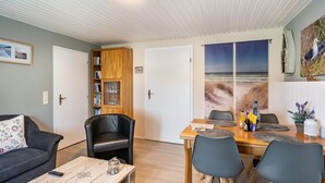 Una sala de estar con un sofá, una mesa de centro, una mesa de comedor con sillas, una estantería y un cuadro de una escena de playa.