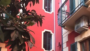 Un edificio rojo con un pequeño balcón, una ventana con contraventanas y una tubería amarilla.