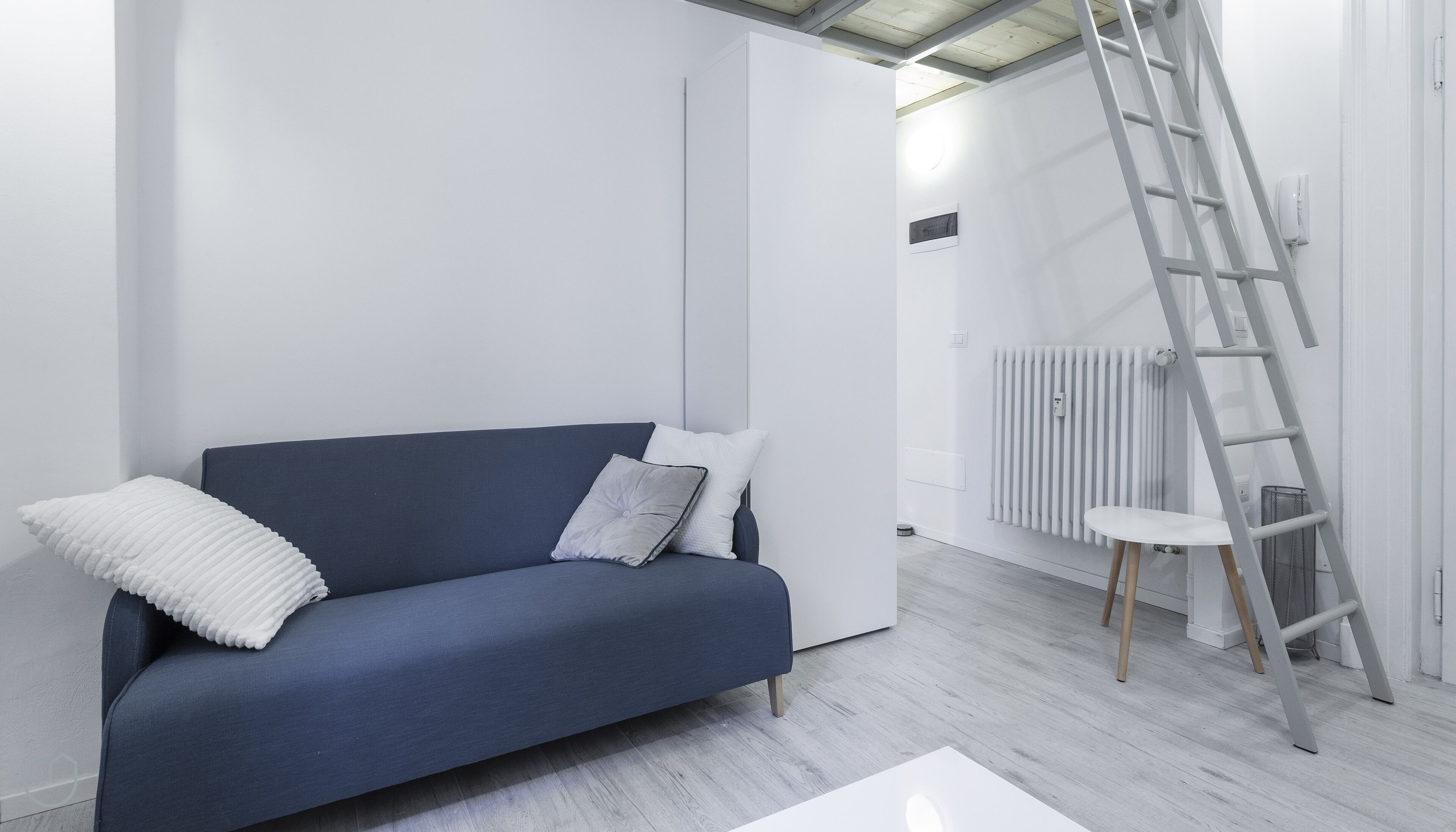 A modern living room with a dark blue sofa, white cushions, a white radiator, and a grey ladder.
