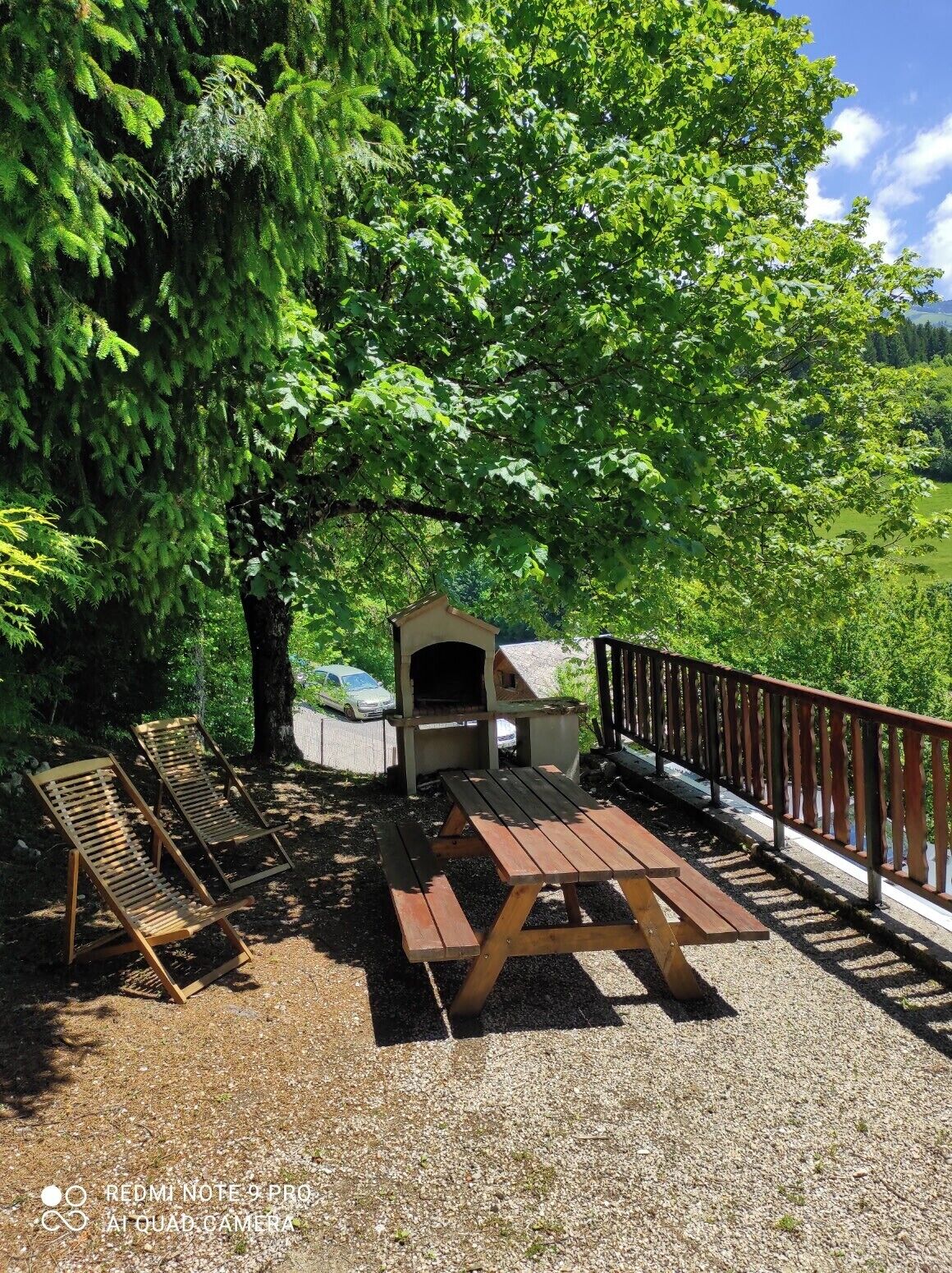 Un área de estar al aire libre con una mesa y bancos de madera, un pequeño horno de ladrillo y una terraza cercada.