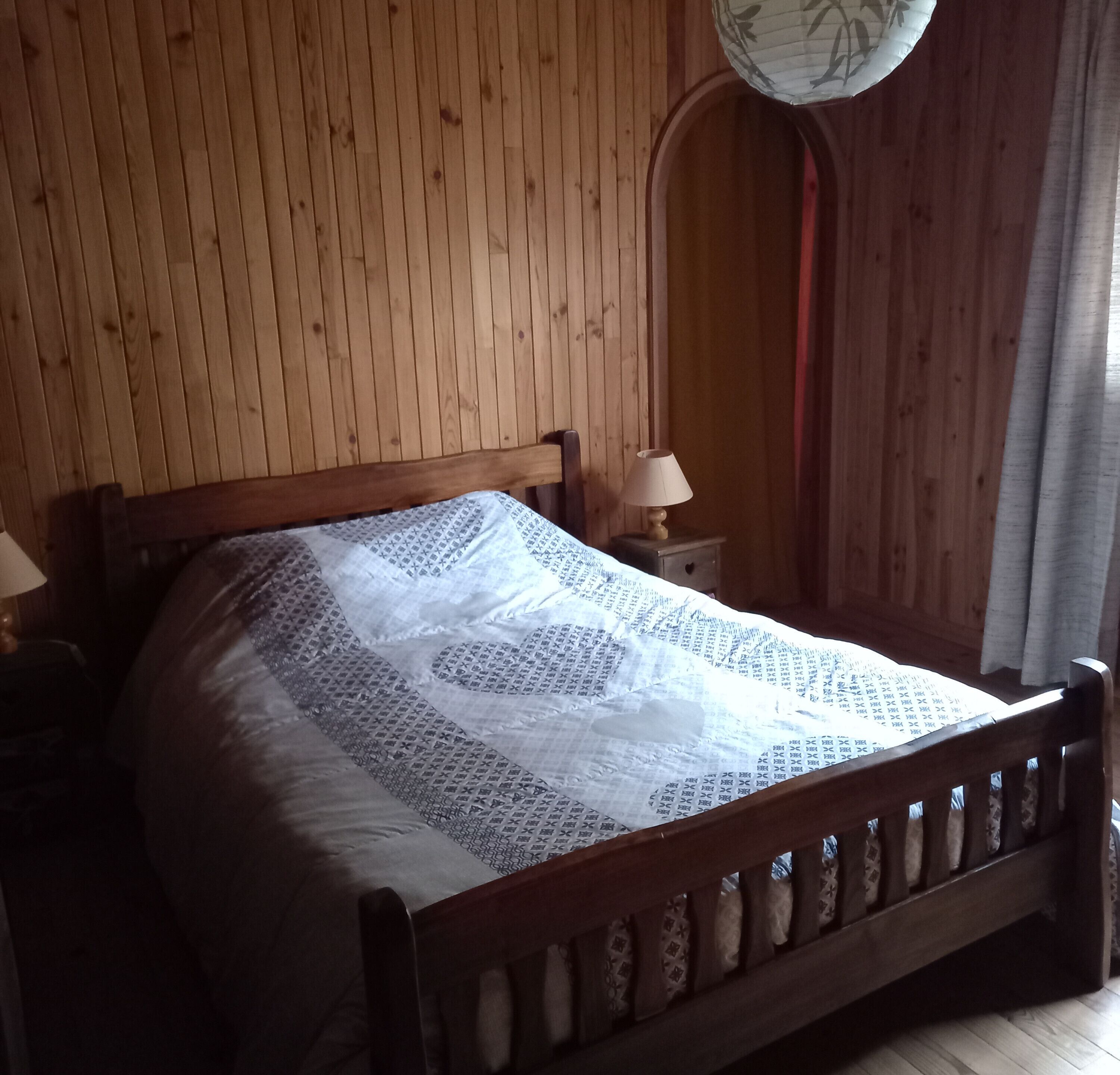 A wooden bed with a patterned bedspread in a room with wooden walls and a window with curtains.