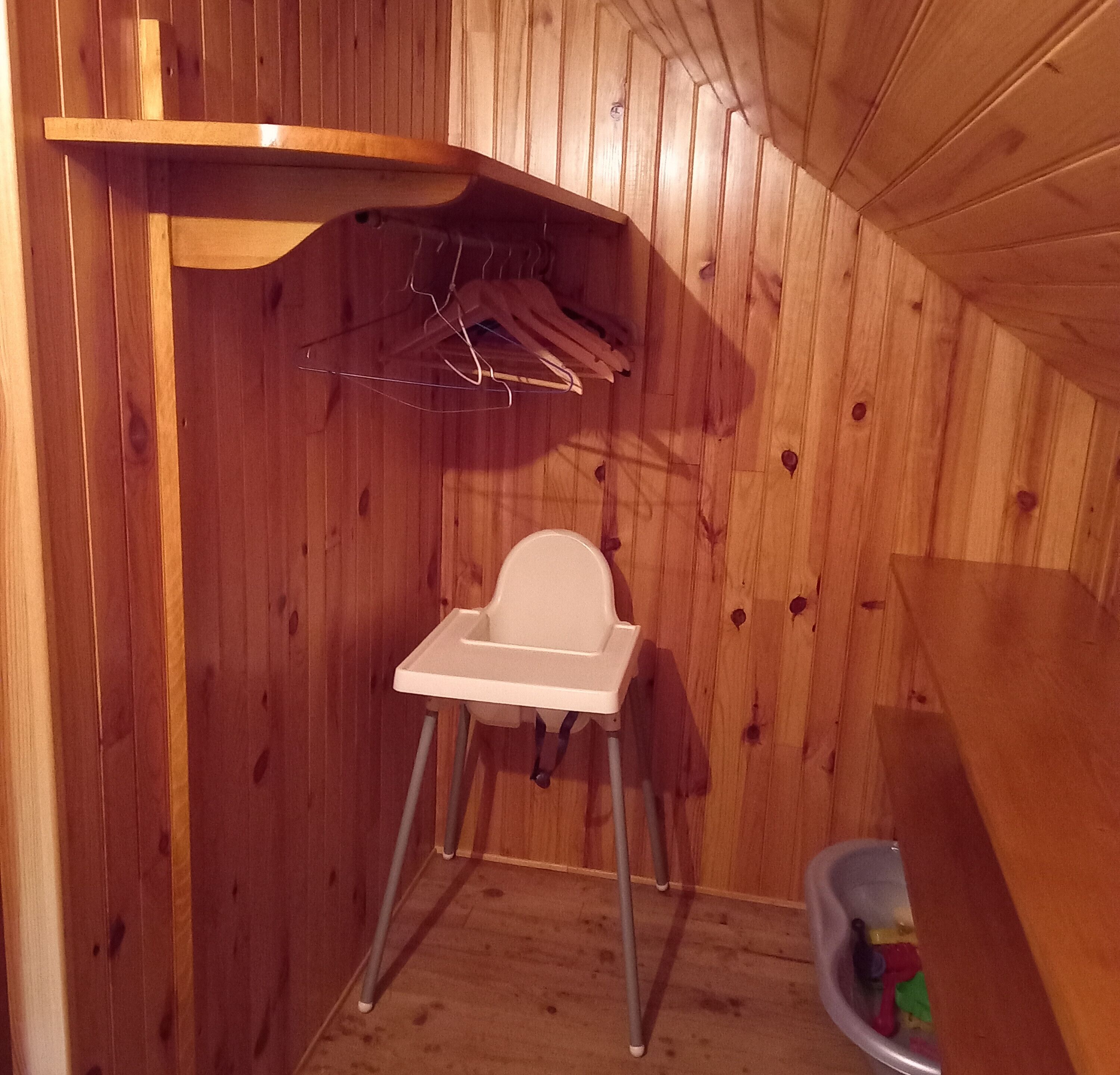 A small wooden room with a white baby high chair, a wooden shelf, and a plastic container with toys.