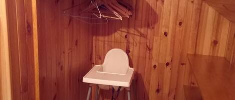 A small wooden room with a white baby high chair, a wooden shelf, and a plastic container with toys.