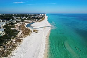 Vista aérea de uma área costeira com uma praia de areia, águas turquesa cristalinas e uma área residencial com casas e um caminho que leva à praia.