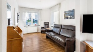 A living room with a brown leather sofa, a wooden piano, a television, and a window with curtains. - Knappstein Holiday Home with 4 Bedrooms, Garden, and Wi-Fi (Möhnesee)