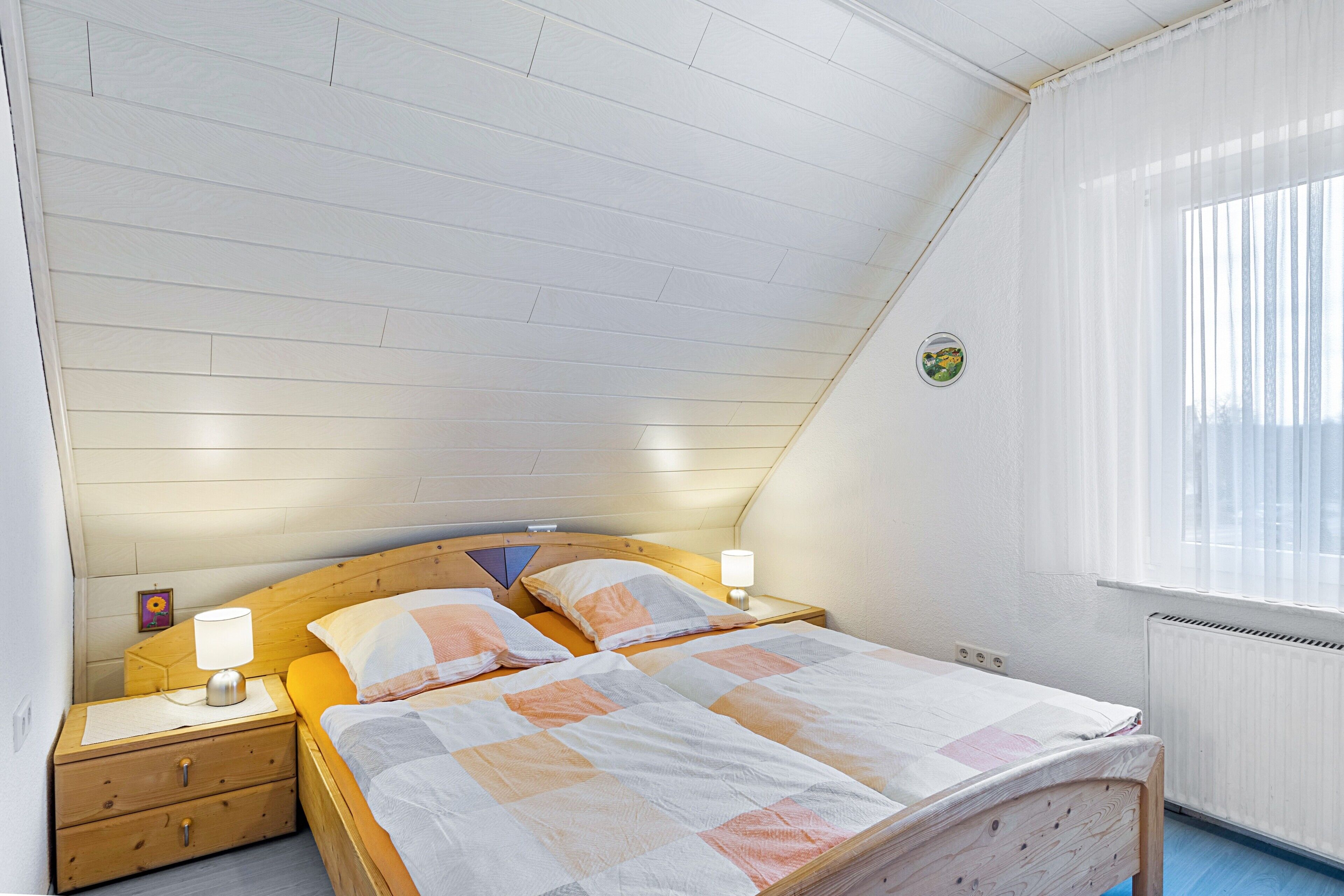 Une chambre avec un plafond en pente, un lit en bois, deux tables de chevet avec des lampes, une fenêtre avec des stores et un radiateur.