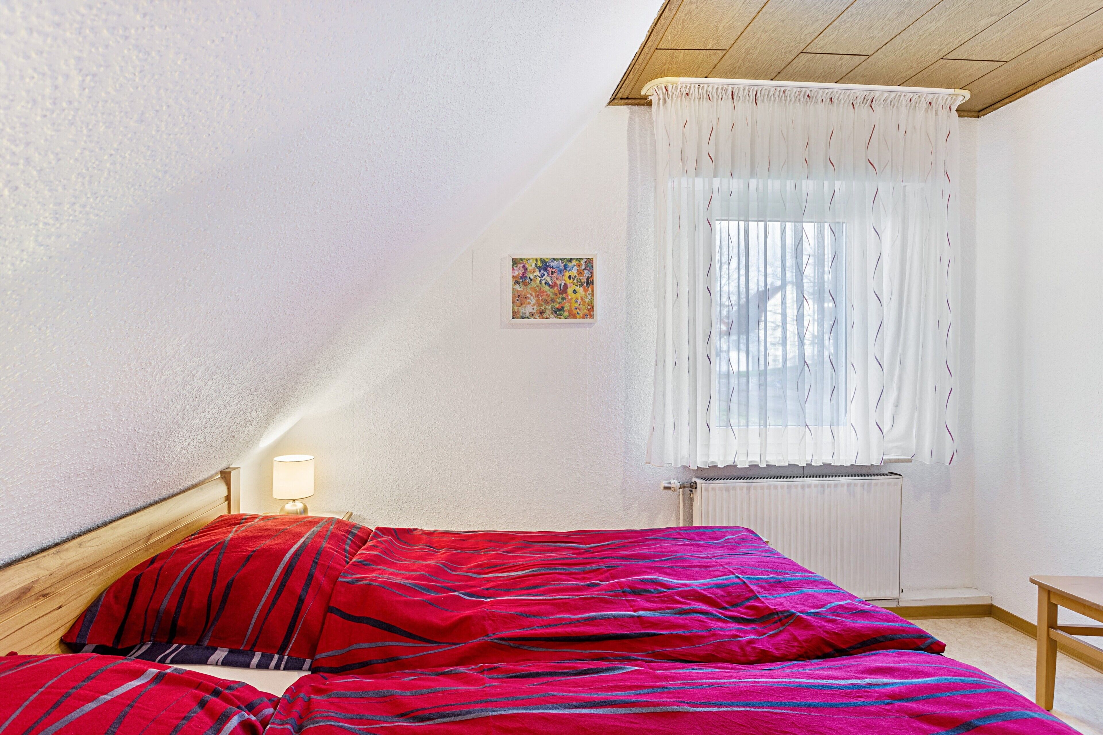 Ein Schlafzimmer mit Bett, Nachttisch, Lampe, Fenster mit Vorhängen und Heizkörper.