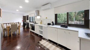 A modern kitchen with white cabinetry, a dining area with a wooden table and chairs, and a large window with a view of greenery. - Casa São Domingos Lousada – Conforto e Vista Deslumbrante para a Vossa Família (Lousada)