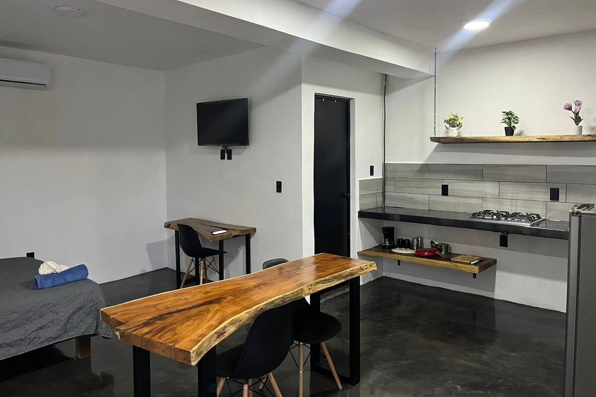 Une cuisine moderne avec une table en bois, des chaises noires, un téléviseur fixé au mur et une porte donnant sur une autre pièce.
