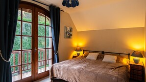 Une chambre avec une grande fenêtre, un lit recouvert d’une couverture marron, deux lampes de chevet et un luminaire au plafond.