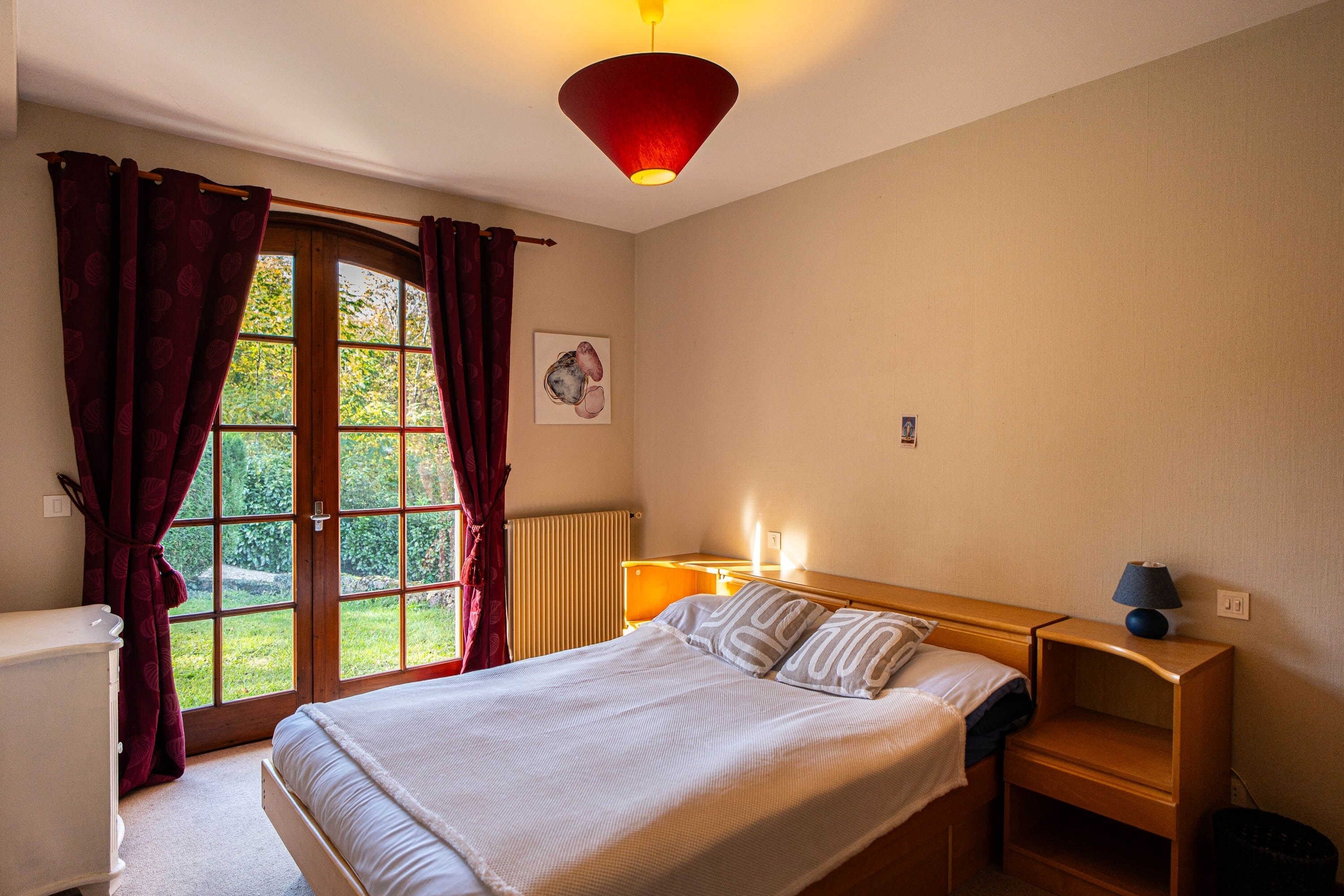 A bedroom with a bed, bedside table, lamp, window with curtains, radiator, and a picture on the wall.