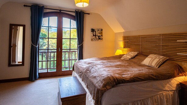 Une chambre à coucher avec un grand lit, un banc, une fenêtre avec des rideaux, un luminaire au plafond et une tête de lit en bois.