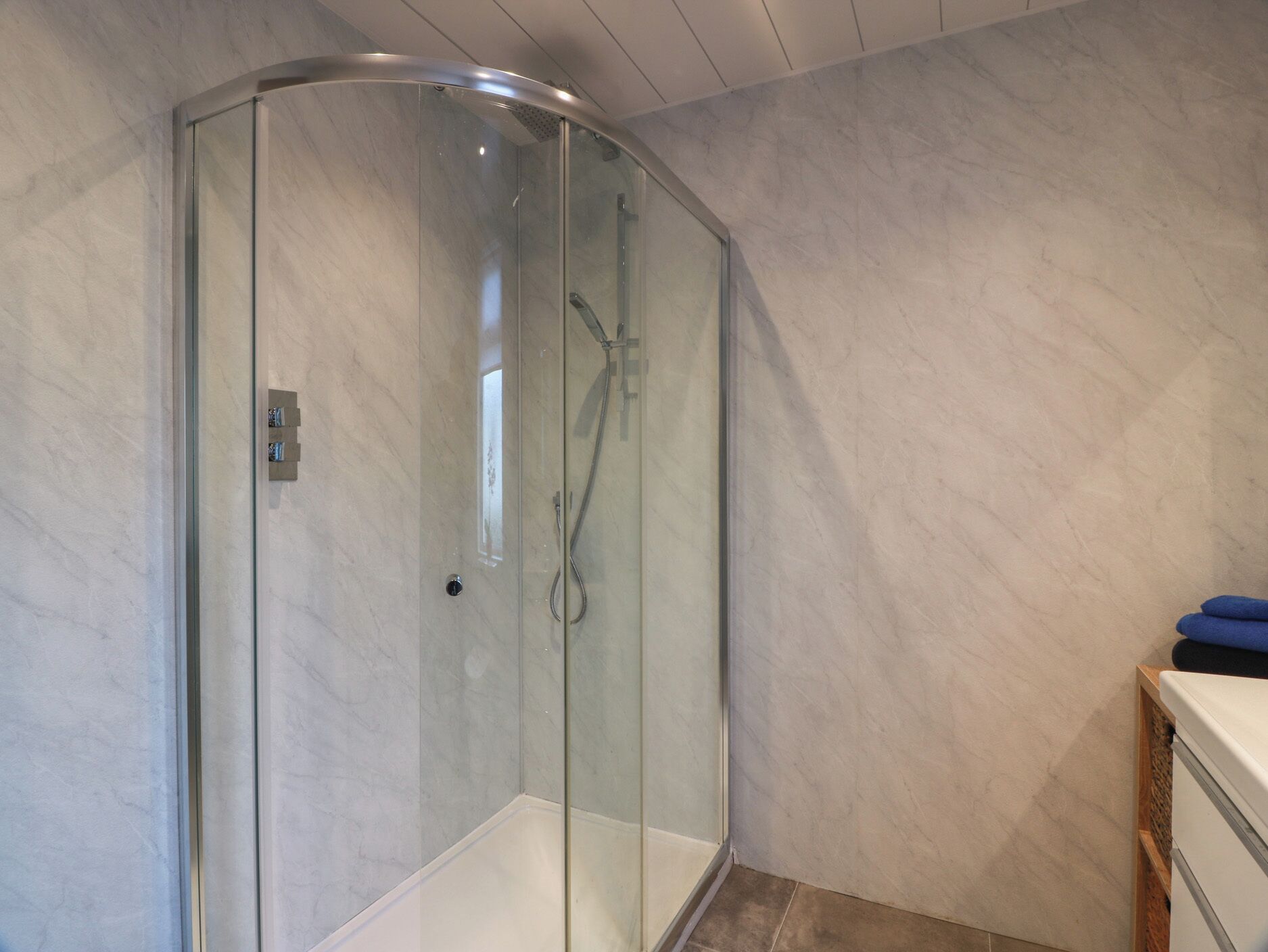 Ducha con cerramiento de vidrio, panel de control, pared de mármol y un mueble de madera con toallas.