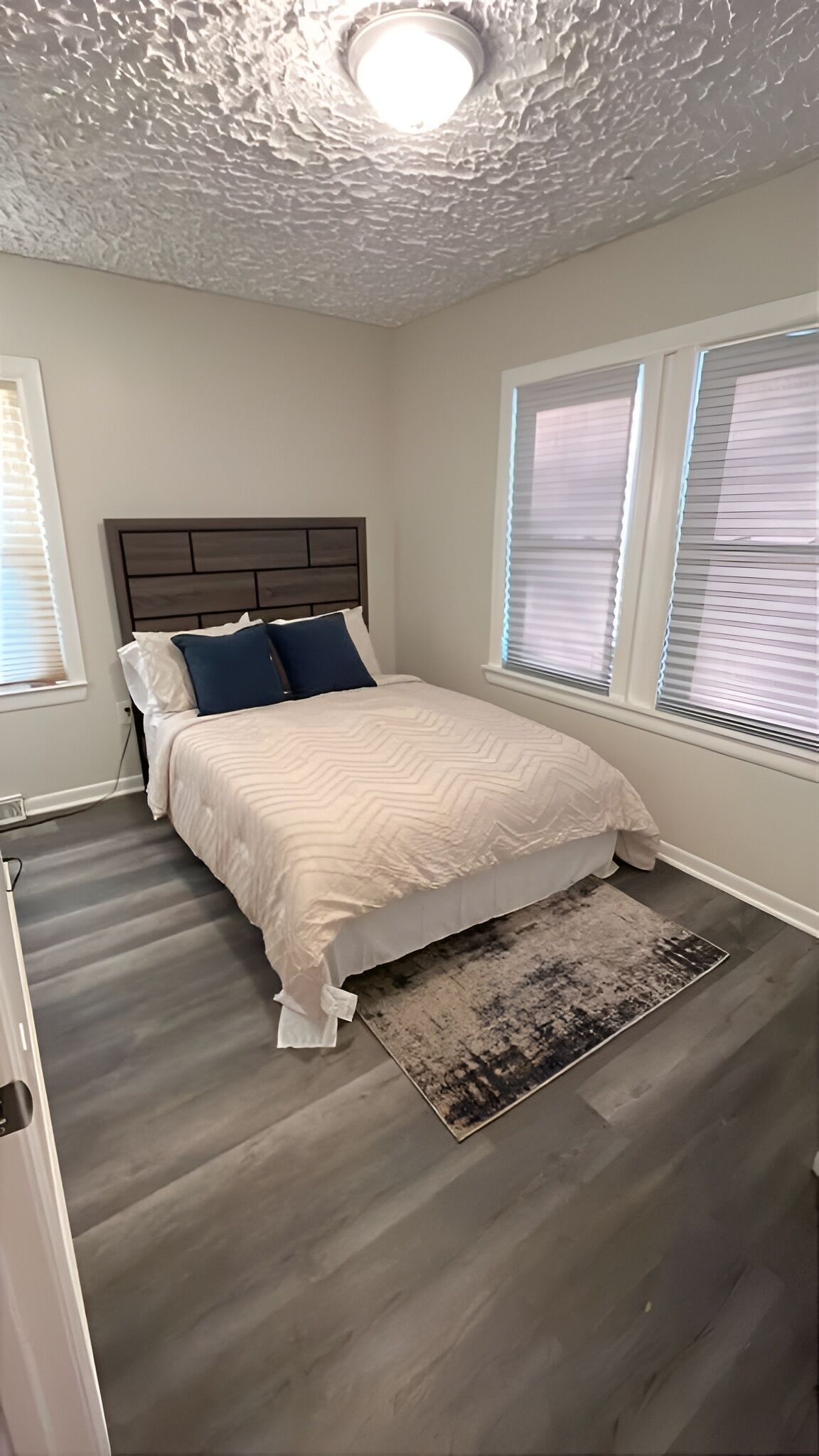 Un dormitorio con cama, cabecera, ventana con persianas y techo con textura.