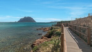 Une promenade en bois mène à une côte rocheuse bordée d’une eau turquoise et limpide, avec une île visible au loin.