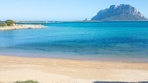Une plage à l’eau d’un bleu limpide, bordée d’un rivage sablonneux, avec une montagne rocheuse imposante qui se dresse à l’arrière-plan.