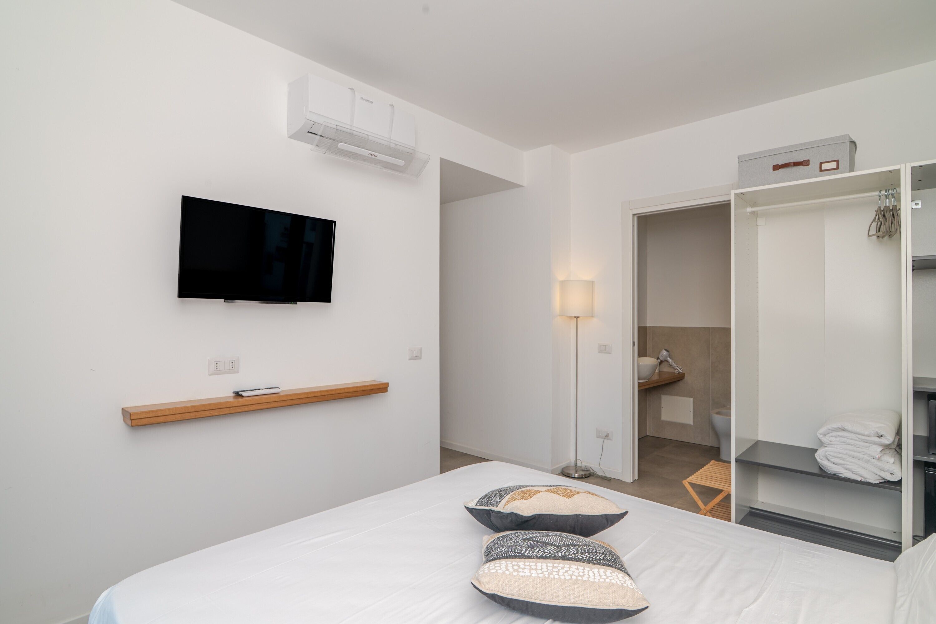 Quarto de hotel moderno com televisão de tela plana, mesa de cabeceira, abajur, guarda-roupa e banheiro visível por uma porta aberta.