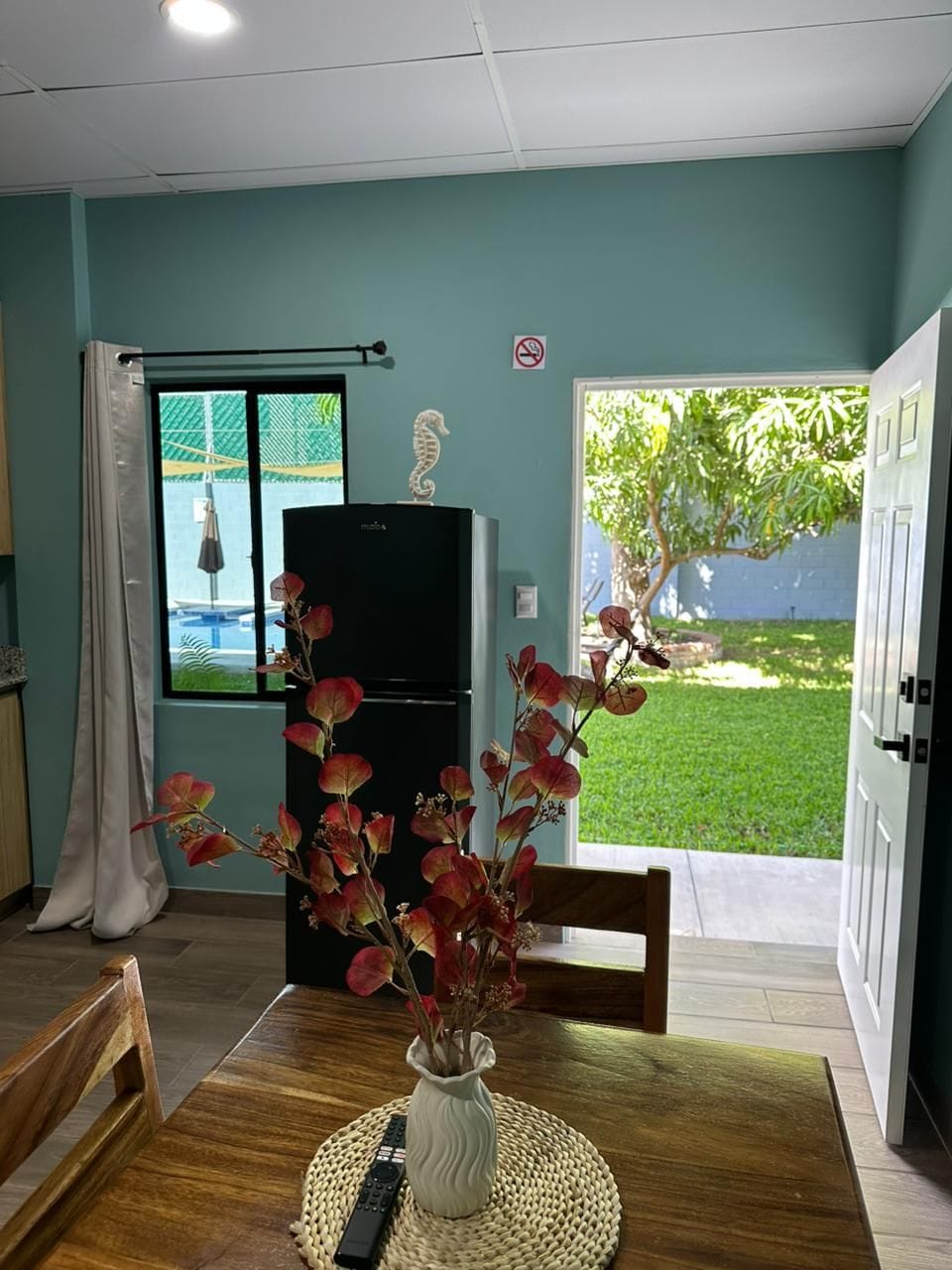 A dining area with a wooden table, a vase with flowers, a black refrigerator, and a view of a backyard with a tree and a pool.