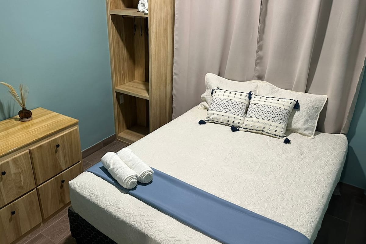 A neatly made bed with a blue bedspread, two pillows with a geometric pattern, and two rolled towels on the side. There is a wooden nightstand with a lamp and a small decorative item. A wooden wardrobe is visible in the background.