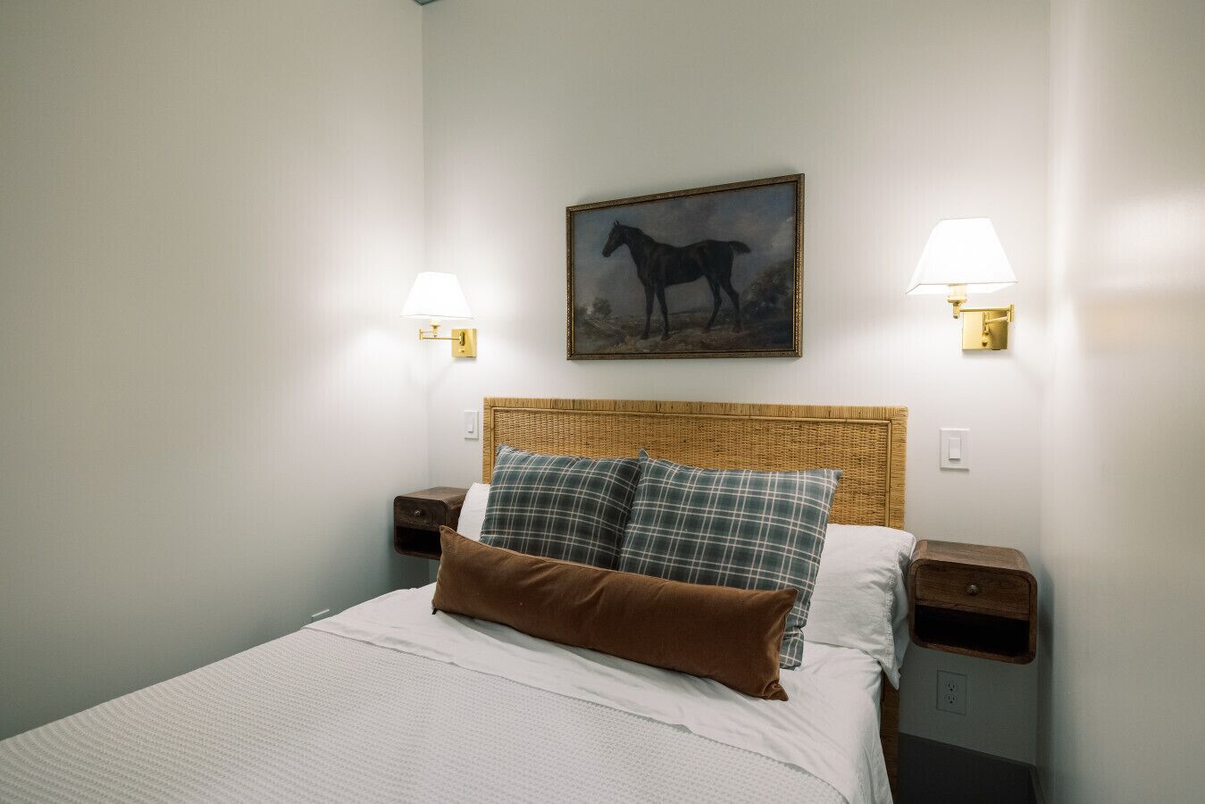 Een netjes opgemaakt bed met een witte deken en twee kussens, een rieten hoofdbord en een schilderij van een paard aan de muur.