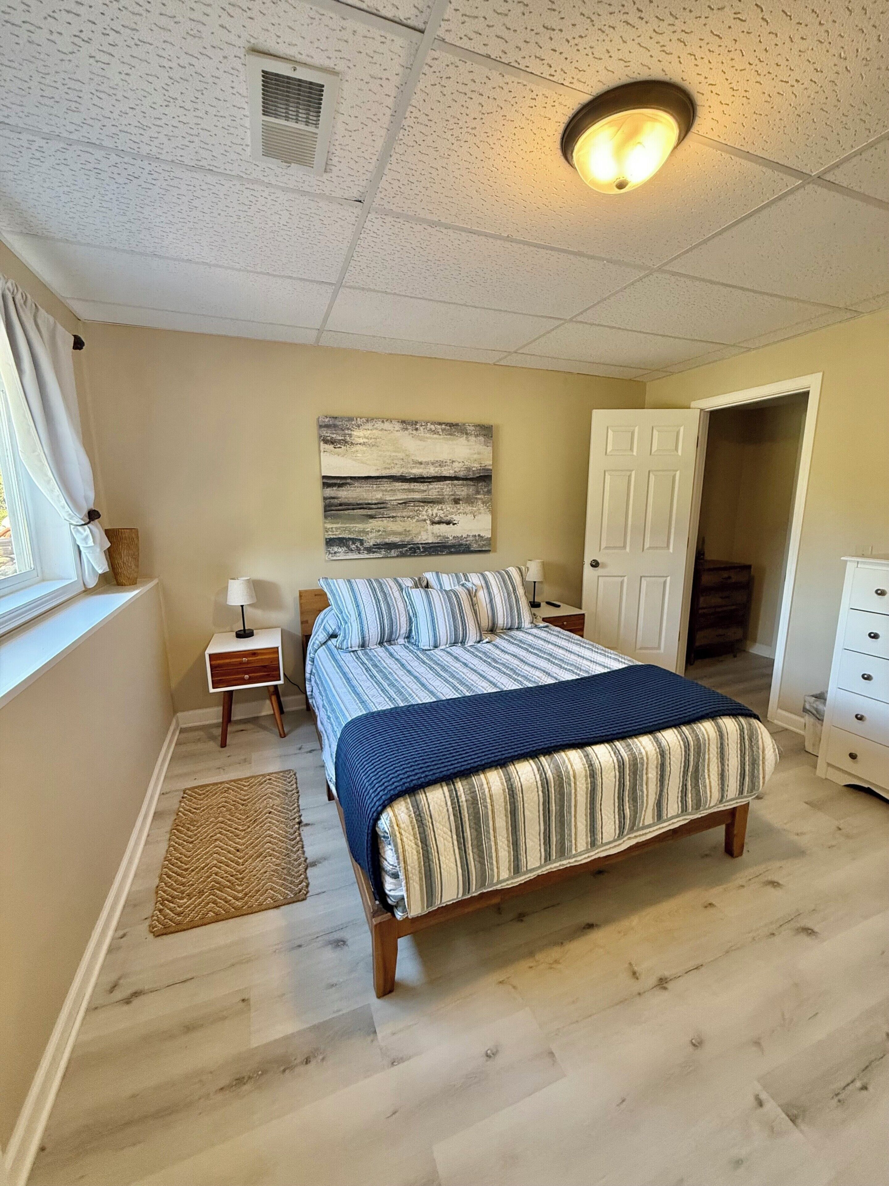 Een slaapkamer met een bed, een nachtkastje, een lamp, een vloerkleed en een deur naar een andere kamer.