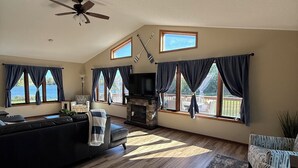A spacious living room with a fireplace, a ceiling fan, and large windows overlooking a body of water.