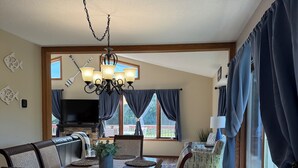 A dining area with a wooden table, chairs, and a chandelier. There is a television in the background and a view of the outdoors through large windows.
