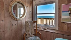 Una habitación de madera con un ventanal grande que da al mar, un espejo redondo y una silla con una manta.
