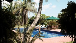 Ein Poolbereich mit Liegestühlen und einem Tisch unter einem grünen Sonnenschirm, umgeben von Palmen und viel Grün.