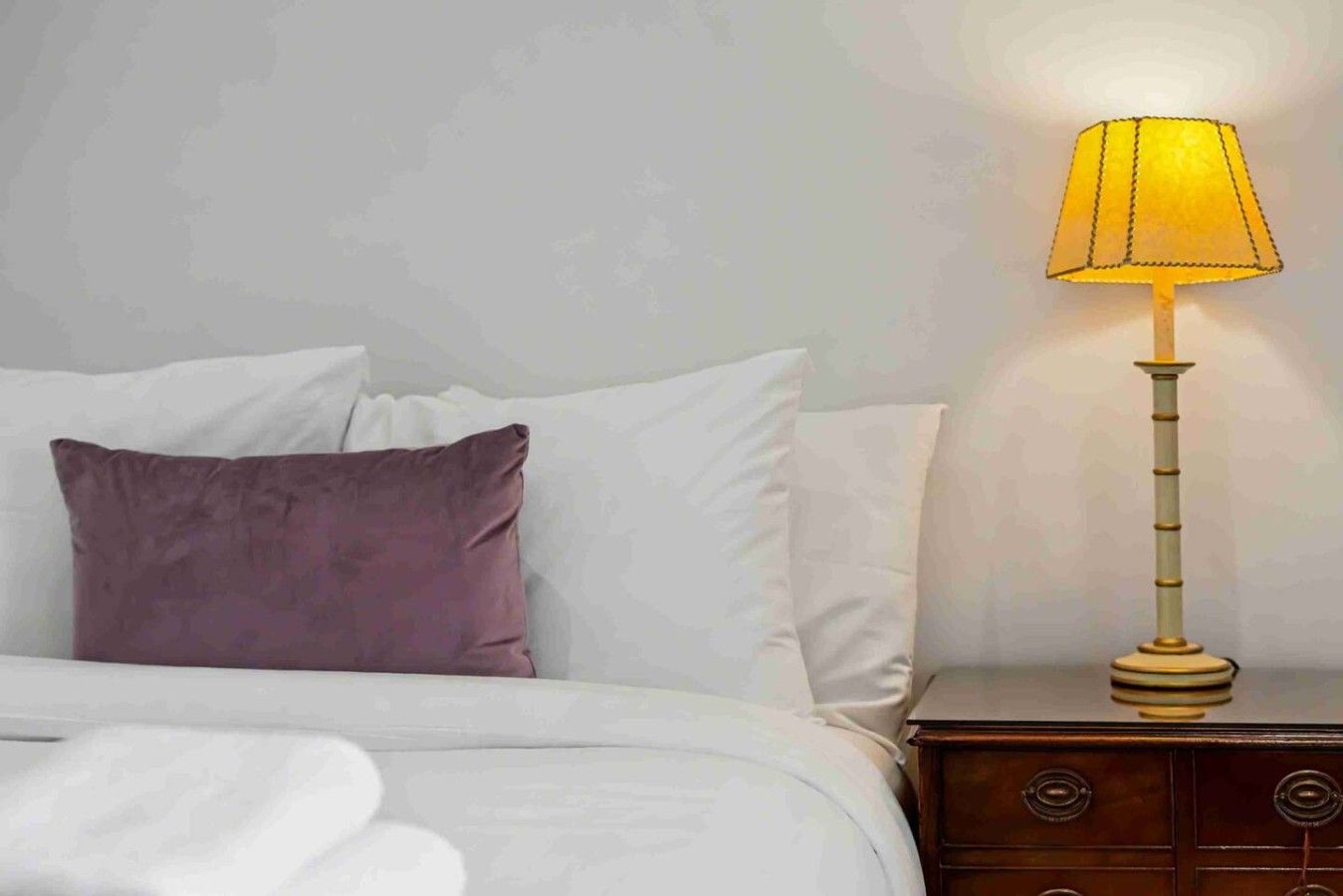 A bed with white linens and a purple pillow, a bedside table with a lamp, and a wooden nightstand.