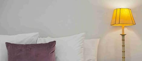 A bed with white linens and a purple pillow, a bedside table with a lamp, and a wooden nightstand.